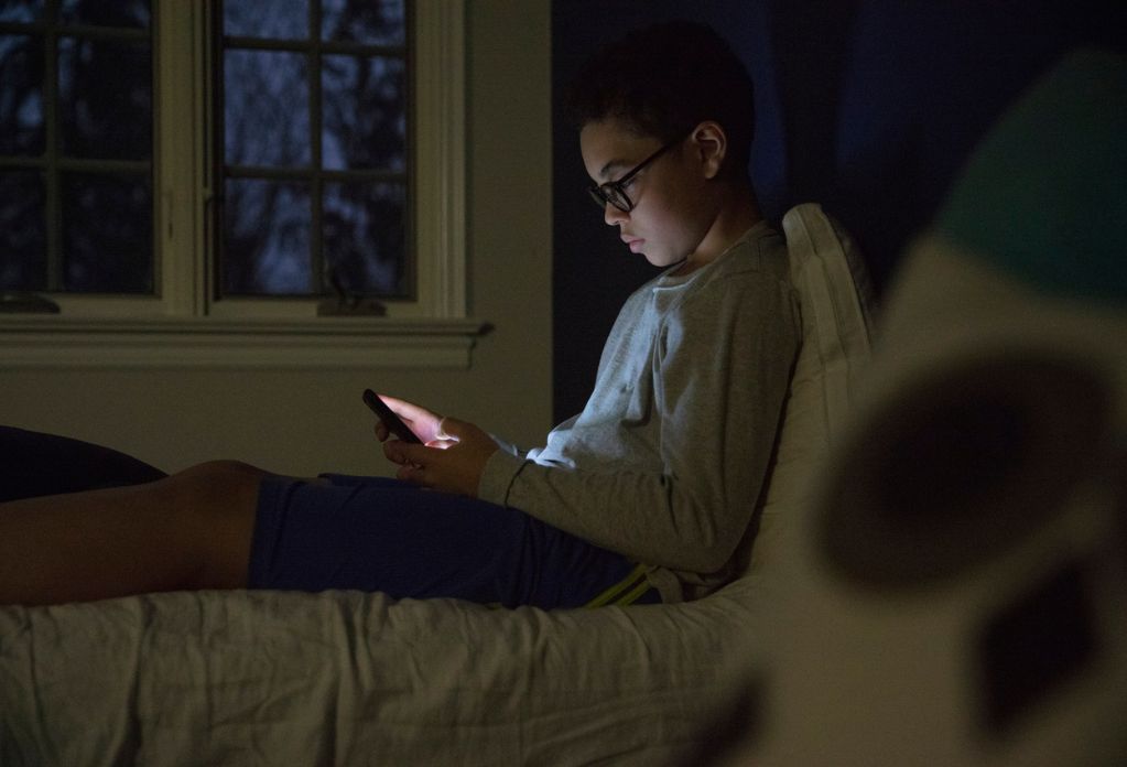 A boy wearing glasses lying on his bed staring at his phone