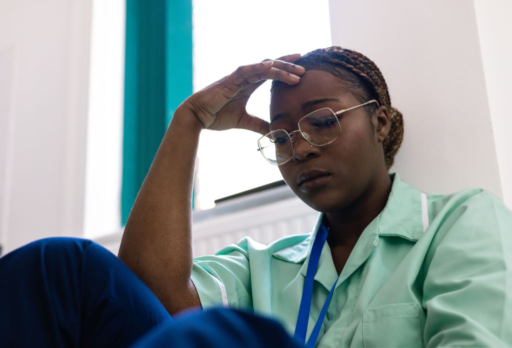 Calls to the modern slavery helpline are at a record high in 2023. Picture is a black women wearing a care worker uniform, looking tired. Hand raised to her head.