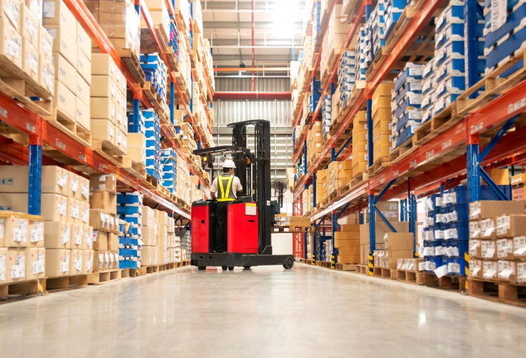 Worker in forklift-truck loading packed goods in huge distribution warehouse with high shelves. Truck unloading goods for logistics company’s modern slavery statement compliance